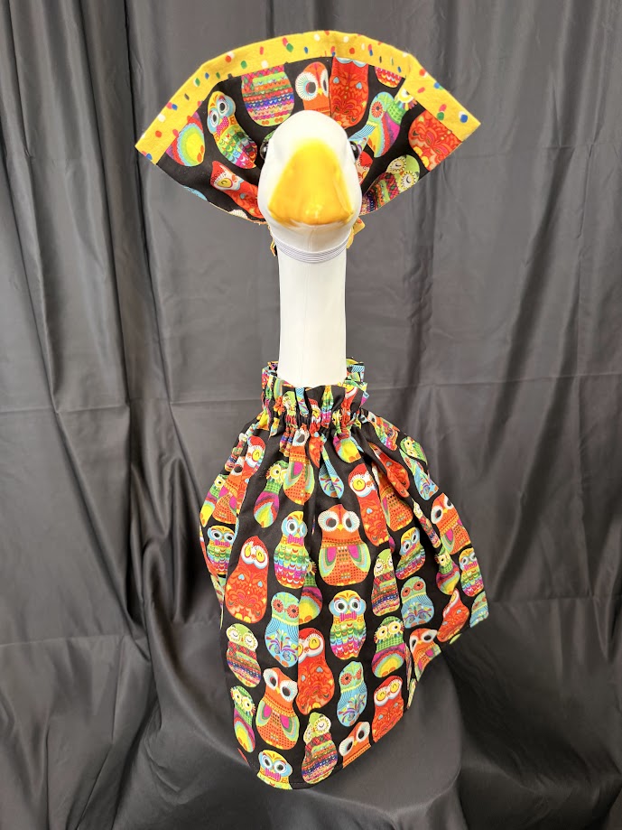 Goose statue wearing black colorful owl print dress and wide yellow/owl reversible hat – front view