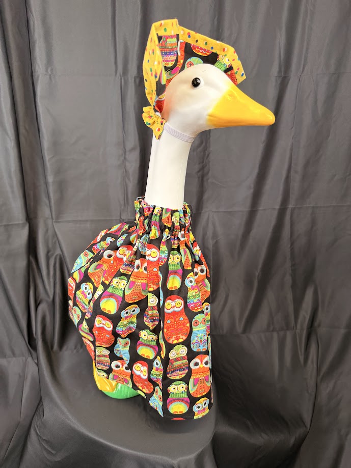Goose statue wearing black colorful owl print dress and yellow/owl reversible hat – angled side view